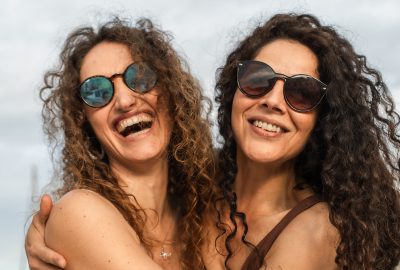 Two cheerful women with curly hair and sunglasses laughing together outdoors. A candid moment of friendship, joy, and happiness, perfect for summer lifestyle and positive emotions.